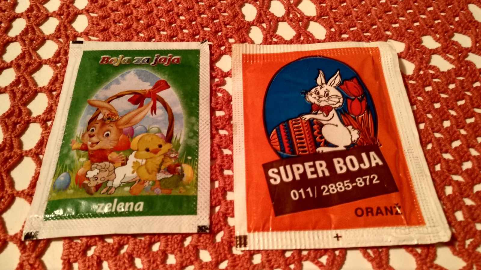 레이스 식탁보 위에 토끼 그림이 그려진 달걀 염색약 봉지 두 개, 'Boja za jaja'와 'SUPER BOJA'라고 적혀 있는 모습